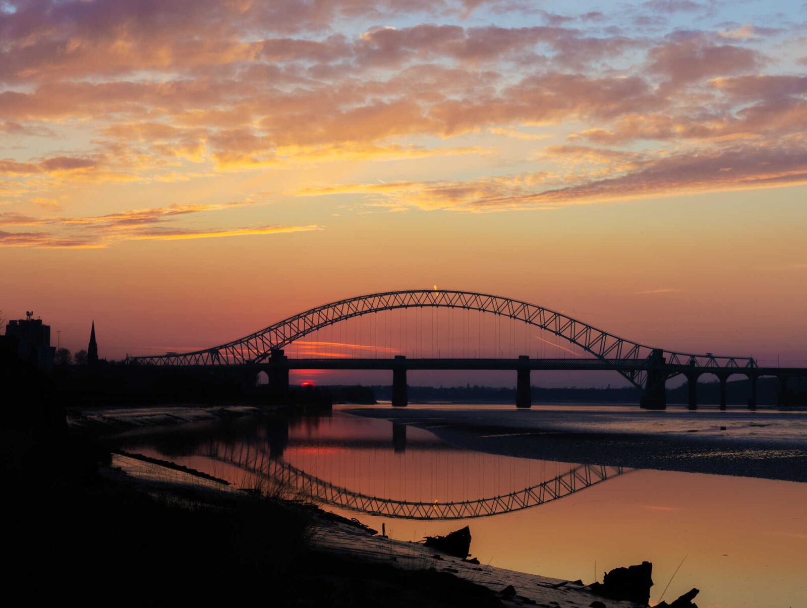 Sunset over the Silver Jubilee Bridge in Runcorn reflected in the calm River Mersey beneath dramatic orange and pink clouds.