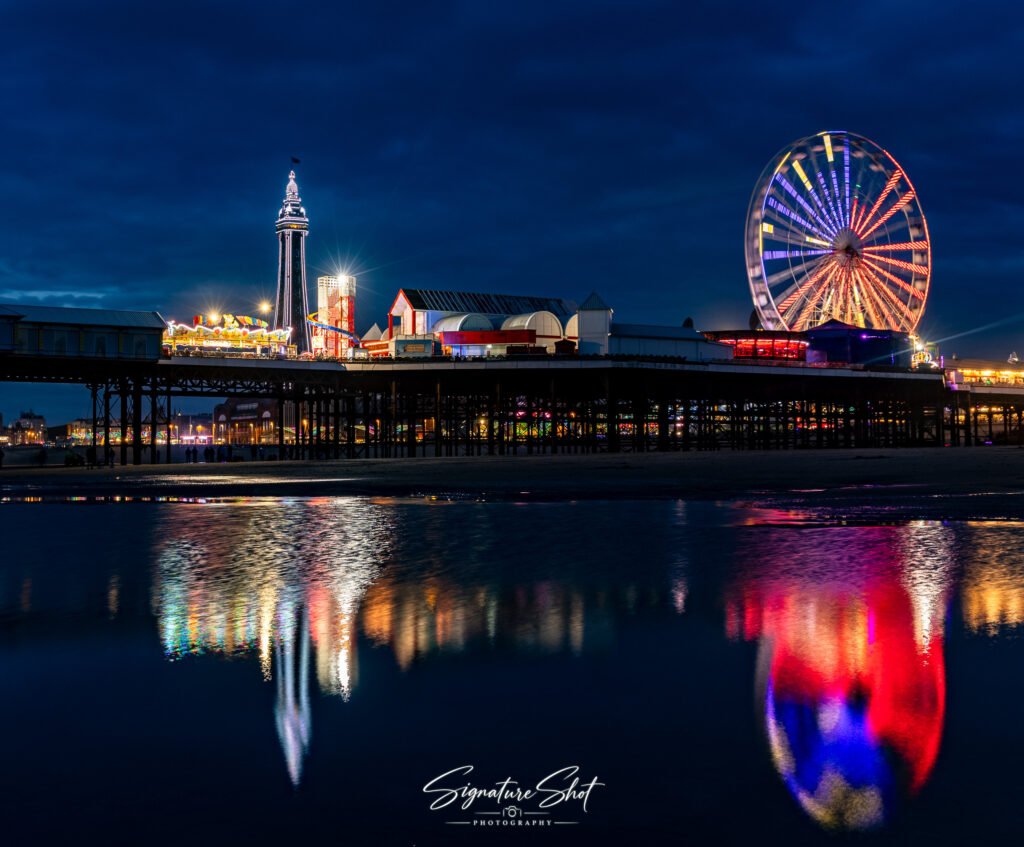Blackpool Tower and Central Pier illuminated at night with the Big Wheel and colourful reflections across the water.