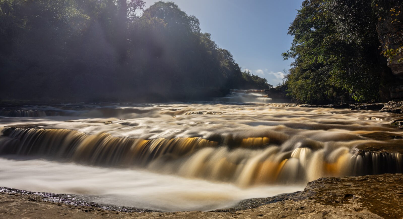 Long-exposure photograph of Aysgarth Lower Falls in the Yorkshire Dales with golden water flowing over limestone ledges beneath surrounding woodland.