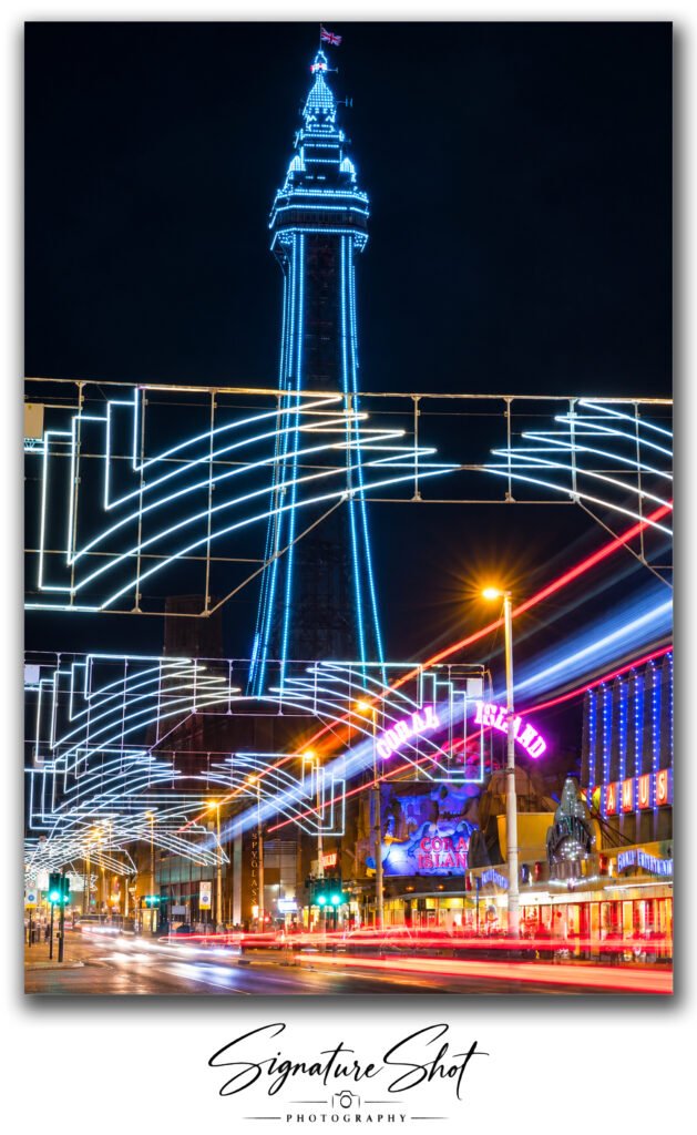 Blackpool Tower illuminated blue at night above Blackpool Promenade, Coral Island and colourful long-exposure traffic light trails.
